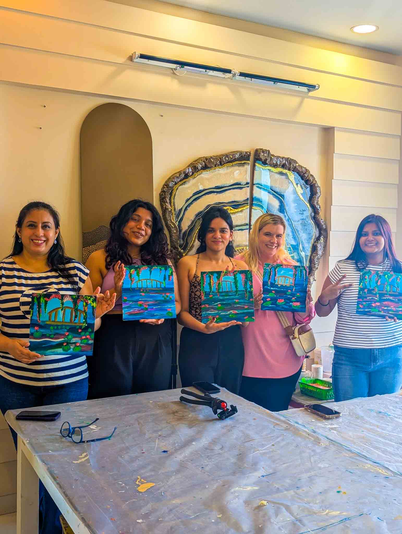 Group of women at a table enthusiastically painting blue scenic canvases during a beginner-friendly Sip & Paint experience.